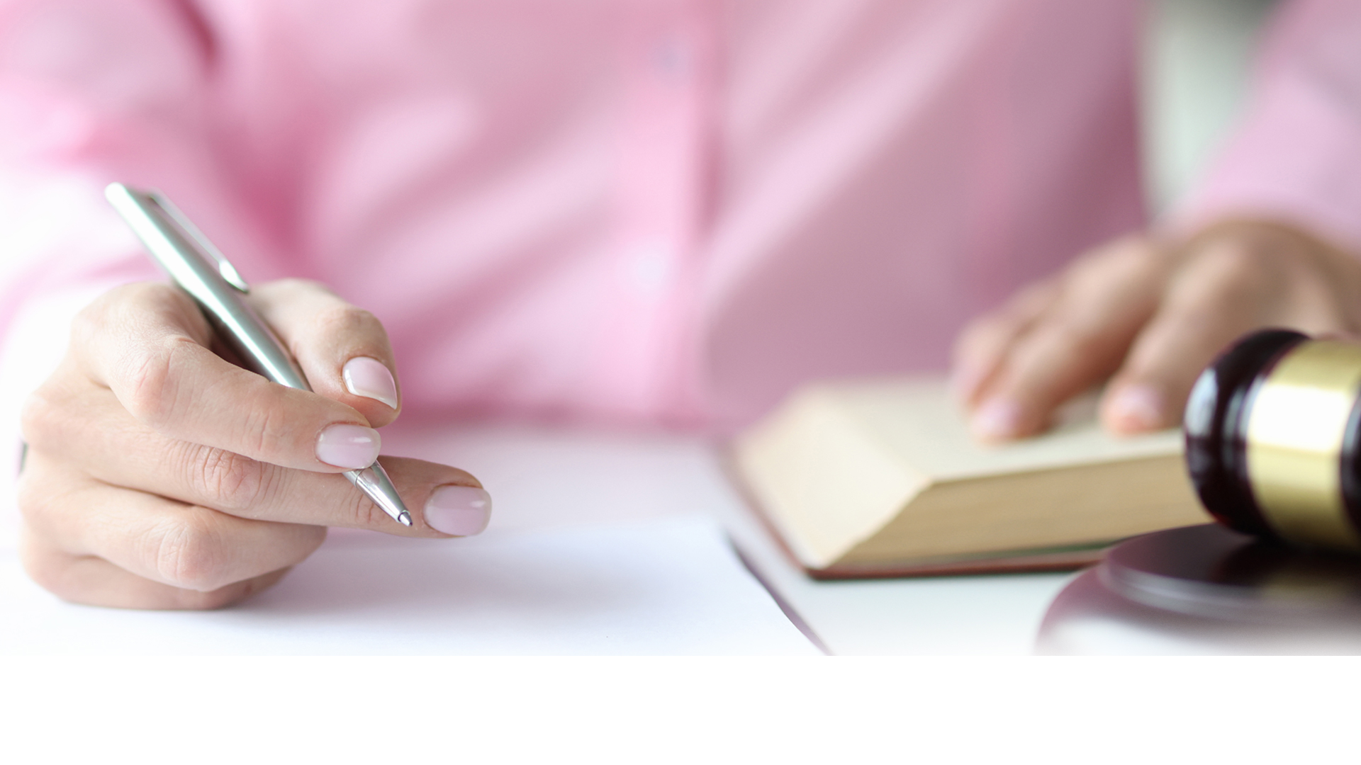 Woman judge writing in documents with ballpoint pen near hammer of courthouse wearing a pink shirt