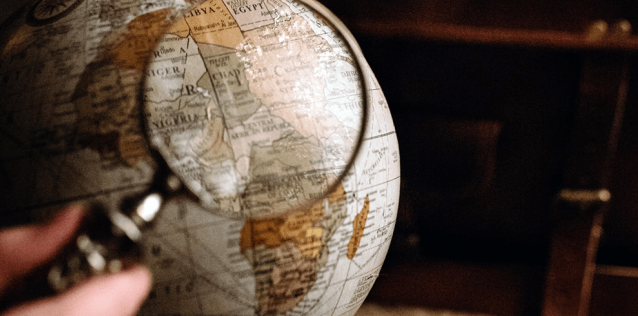 A hand holding a magnifying glass in front of an earth globe with an old leather breafcase in the background.